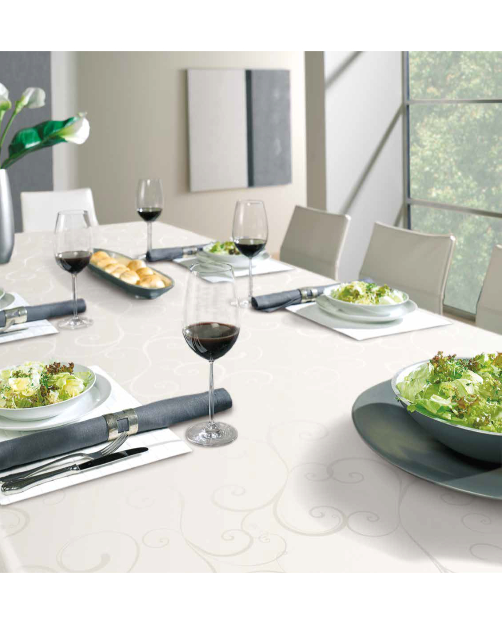 Moderni ruokapöytä, jossa on Pyöreä vahakangasliina Ø140cm, valkoisia salaattilautasia, punaviinillä täytettyjä viinilaseja, harmaita lautasliinoja ja pitkä leipälautanen valoisassa huoneessa, jossa on valkoiset tuolit ja suuret ikkunat.