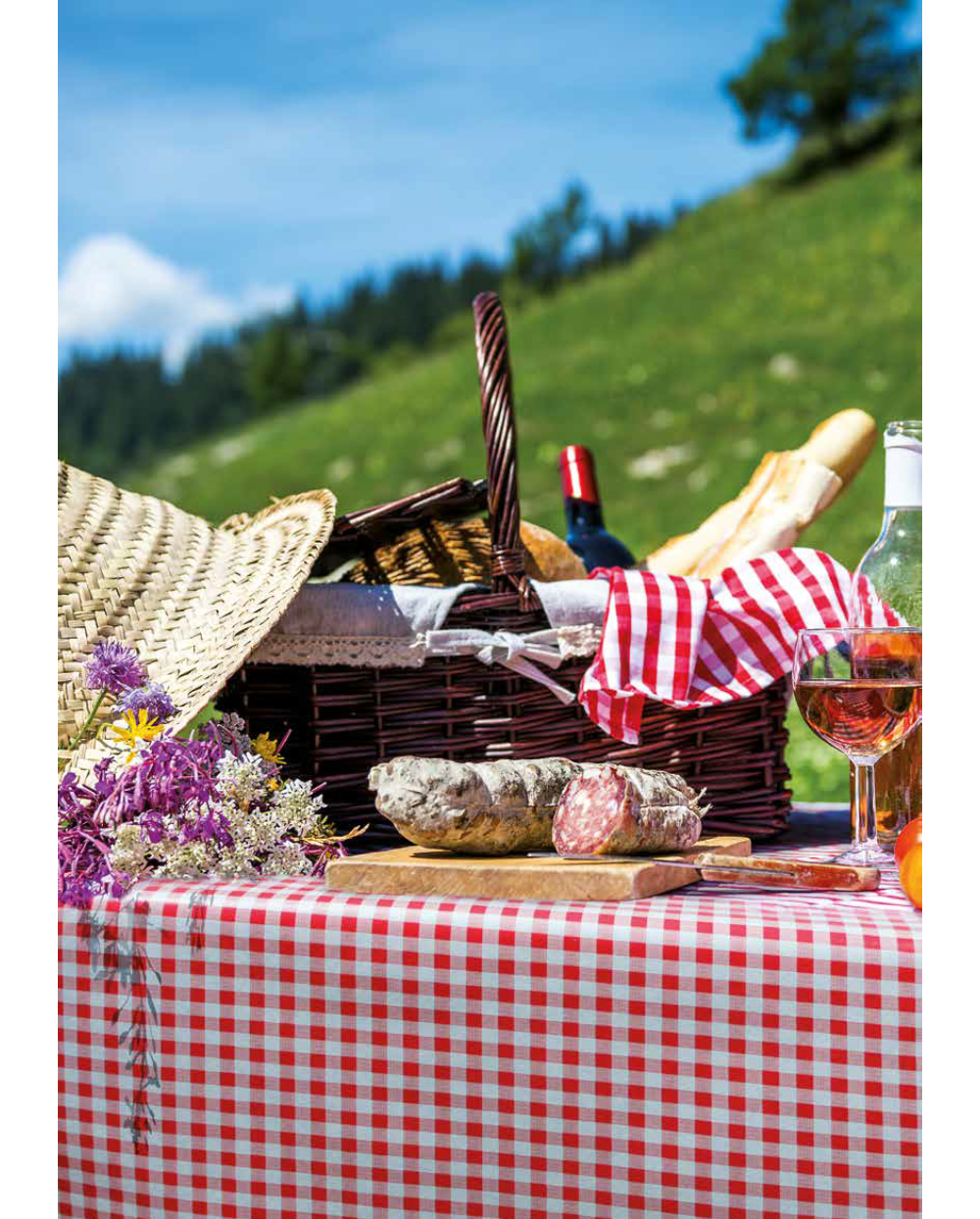 Pyöreä vahakangasliina Ø140cm pöytäliina, pajukori, patonki, viini, makkarat, villikukat ja olkihattu, jotka on järjestetty ulkona nurmikolla sinisen taivaan alla.