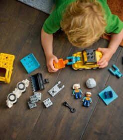 Lapsi leikkii lattialla erilaisilla rakenteluaiheisilla LEGO Duplo -palikoilla, joihin kuuluu kuorma-auto, hahmot ja rakennuspalikat.