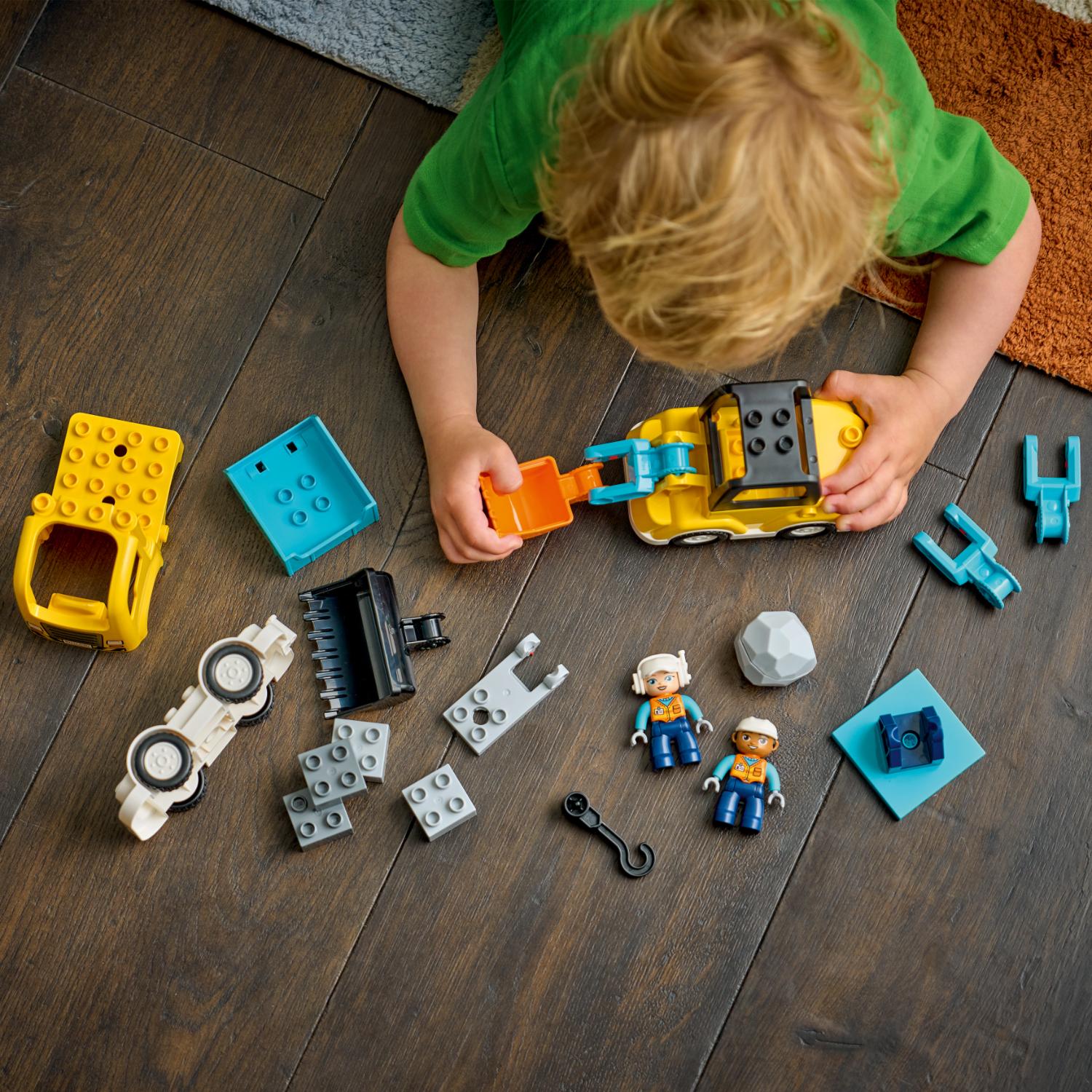 Lapsi leikkii lattialla erilaisilla rakenteluaiheisilla LEGO Duplo -palikoilla, joihin kuuluu kuorma-auto, hahmot ja rakennuspalikat.