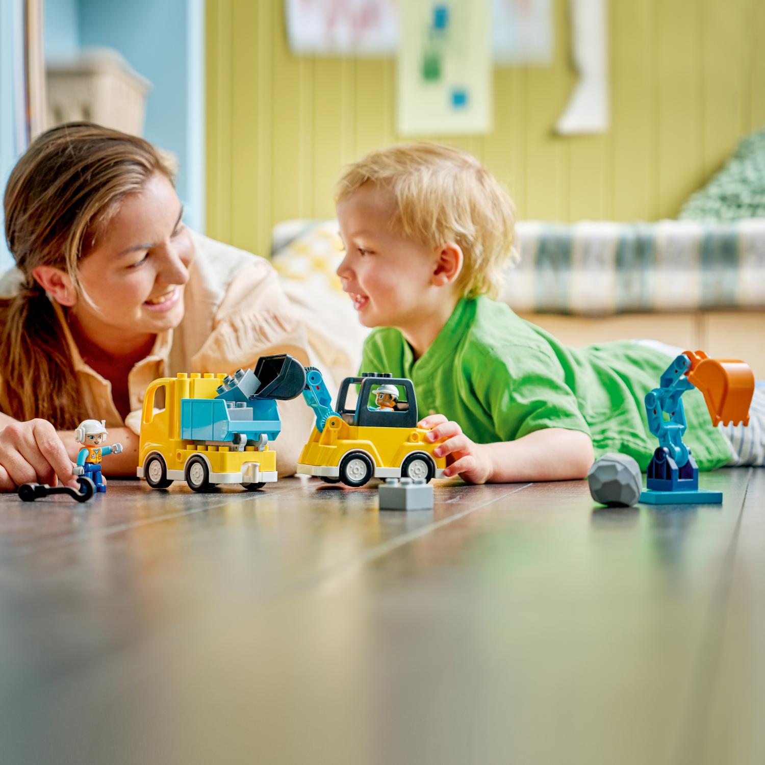 Nainen ja pieni poika leikkivät yhdessä lattialla keltaisilla LEGO-rakennusajoneuvoilla ja -hahmoilla.