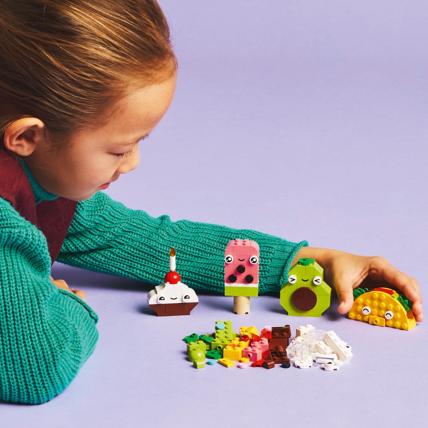 Lapsi ojentaa kätensä LEGO-tacoa kohti, ja hänen edessään on rivi muita ruoan muotoisia LEGO-luomuksia, kuten cupcake, jäätelöpuikko ja avokado.
