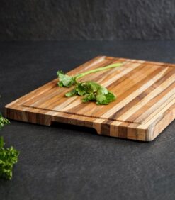 Leikkuulauta, jossa on 45 x 30 cm:n kokoinen tiikkipuu ja jonka päällä on korianterin oksa, seisoo tummalla työtasolla, ja vasemmassa alakulmassa näkyy hieman persiljaa.