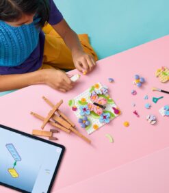 Henkilö, joka kokoaa kukanmuotoista LEGO-mosaiikkia vaaleanpunaisella pöydällä tabletin vieressä, jonka näytöllä näkyy kokoamisohjeet.