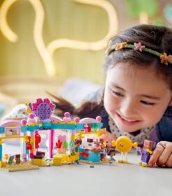 Nuori tyttö hymyilee leikkiessään värikkäällä Lego Friends -rakennussarjalla, jossa on kauppa ja pienoishahmot.