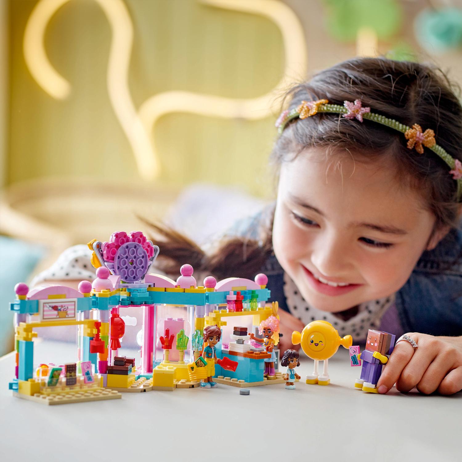 Nuori tyttö hymyilee leikkiessään värikkäällä Lego Friends -rakennussarjalla, jossa on kauppa ja pienoishahmot.