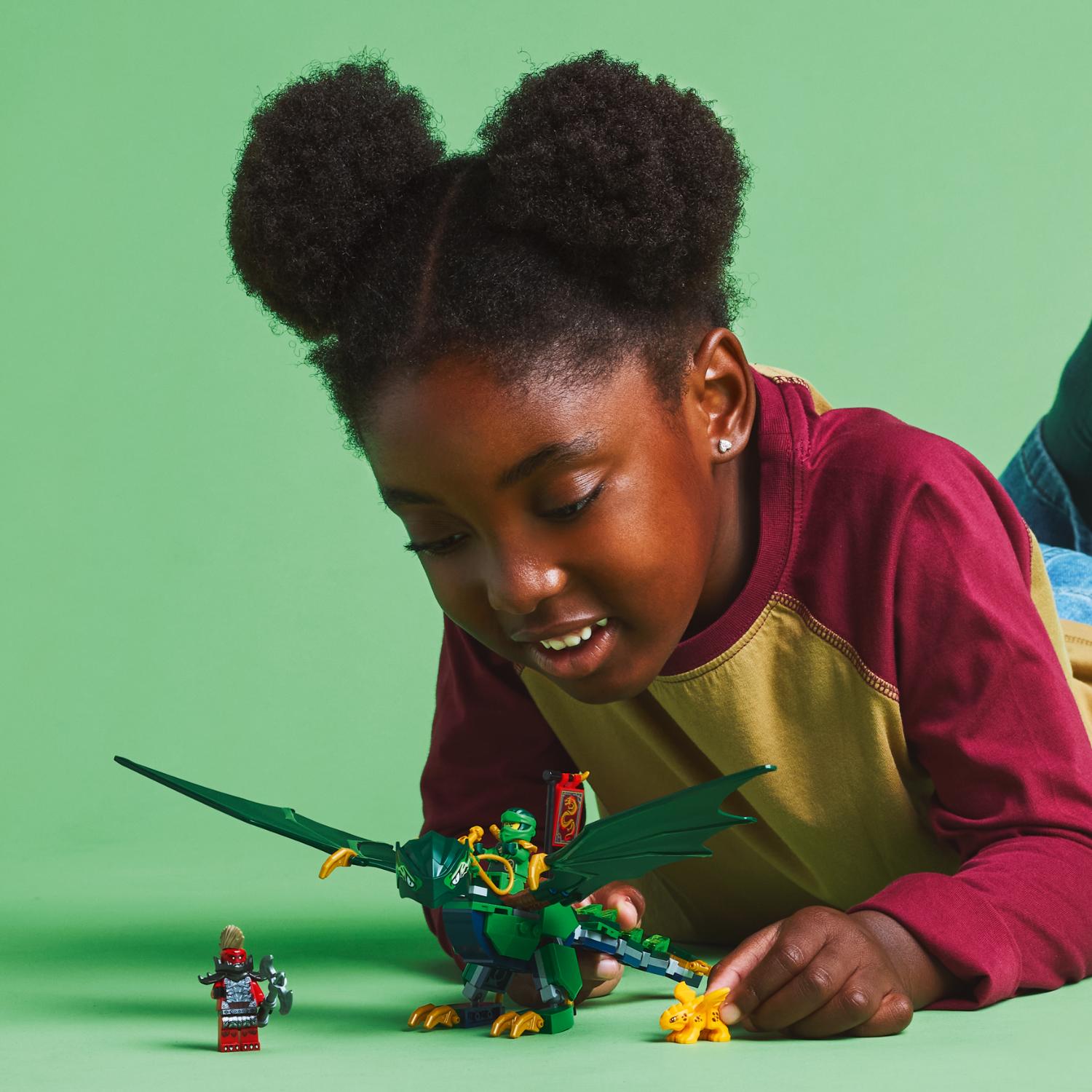 Nuori tyttö leikkii vihreällä taustalla vihreällä Lego-lohikäärmeellä, Lego-minihahmolla ja pienellä kultaisella olennolla.