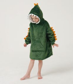 Lapsi, jolla on yllään vihreä, dinosauruksia esittävä huppupeitto, jossa on oransseja piikkejä, seisoo paljain jaloin valkoisella taustalla.