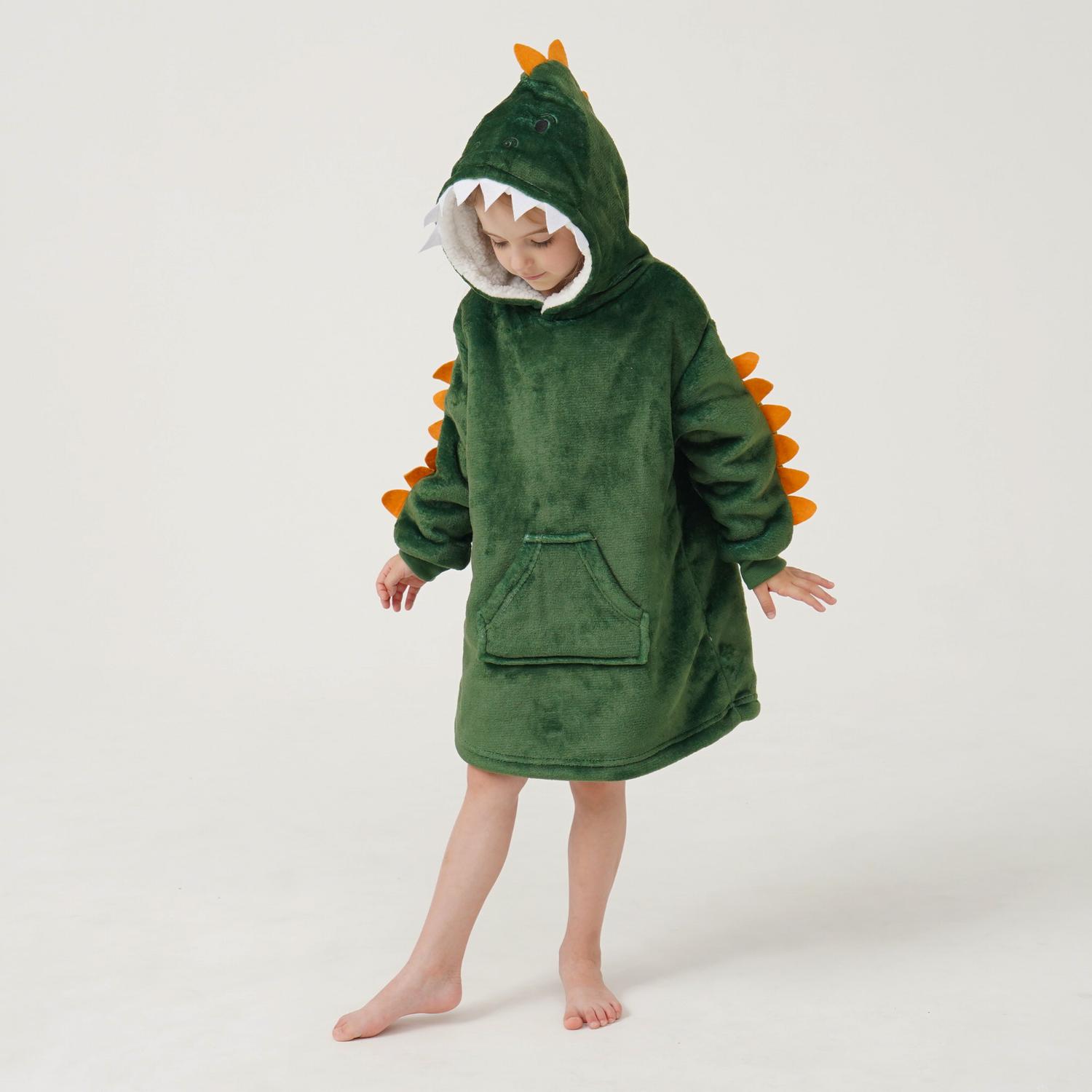 Lapsi, jolla on yllään vihreä, dinosauruksia esittävä huppupeitto, jossa on oransseja piikkejä, seisoo paljain jaloin valkoisella taustalla.