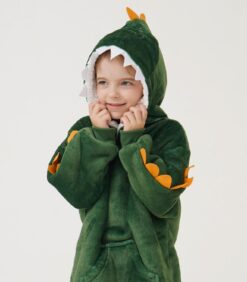 Pieni lapsi hymyilee pukeutuneena vihreään dinosauruspukuun, jonka hupussa on hampaat, silmät ja oranssit piikit, ja seisoo vaalealla, yksivärisellä taustalla.