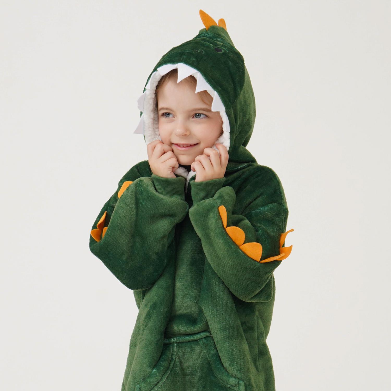 Pieni lapsi hymyilee pukeutuneena vihreään dinosauruspukuun, jonka hupussa on hampaat, silmät ja oranssit piikit, ja seisoo vaalealla, yksivärisellä taustalla.