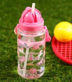 Lasten juomapullo yksisarvinen, kirkas vesipullo, jossa on vaaleanpunainen kansi ja hihna, jossa on yksisarvisen ja prinsessan kuvioita, on sijoitettu vihreälle tekonurmelle keltaisen tennispallon ja punaisen mailan viereen.