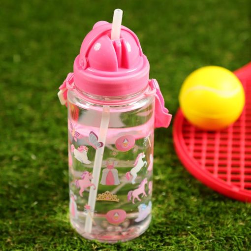 Lasten juomapullo yksisarvinen, kirkas vesipullo, jossa on vaaleanpunainen kansi ja hihna, jossa on yksisarvisen ja prinsessan kuvioita, on sijoitettu vihreälle tekonurmelle keltaisen tennispallon ja punaisen mailan viereen.