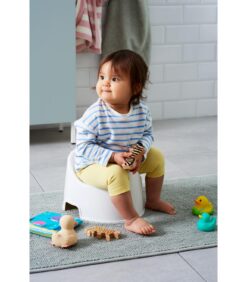 Pikkulapsi istuu kylpyhuoneessa valkoisella potalla ja pitelee kädessään leluseepraa. Leluja, kirja ja kumiankat ovat hajallaan lähellä olevalla harmaalla matolla. Lapsella on päällään raidallinen paita ja keltaiset housut.