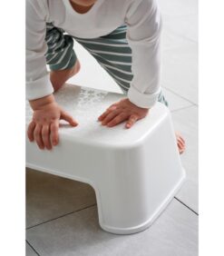 Pieni lapsi, jolla on raidalliset housut ja valkoinen paita, kiipeää laattalattialle sijoitettuun valkoiseen muoviseen jakkaraan käyttäen molempia käsiään tukena.
