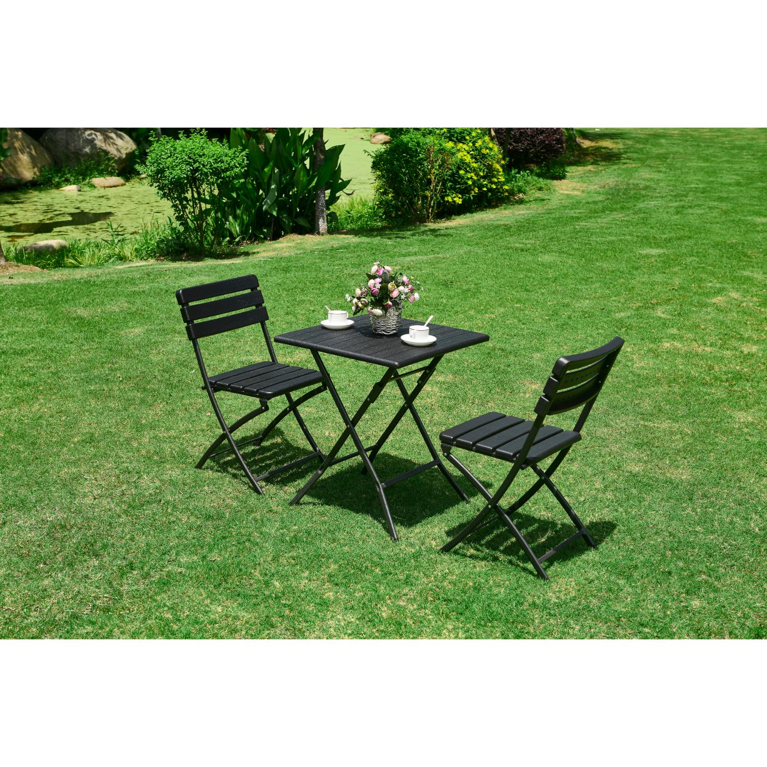 Pieni musta taittopöytä ja kaksi mustaa taittotuolia pöydän ääressä. Pöytä ja tuolit ovat nurmikolla.