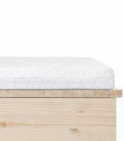Lähikuva Vauvanpatja 60x120x5 original uusi kuosi -patjasta, jossa on valkoinen päällinen, jossa on hienovarainen kolmiokuvio, ja joka on asetettu vaaleanpuiseen puiseen sängynrunkoon valkoista taustaa vasten.