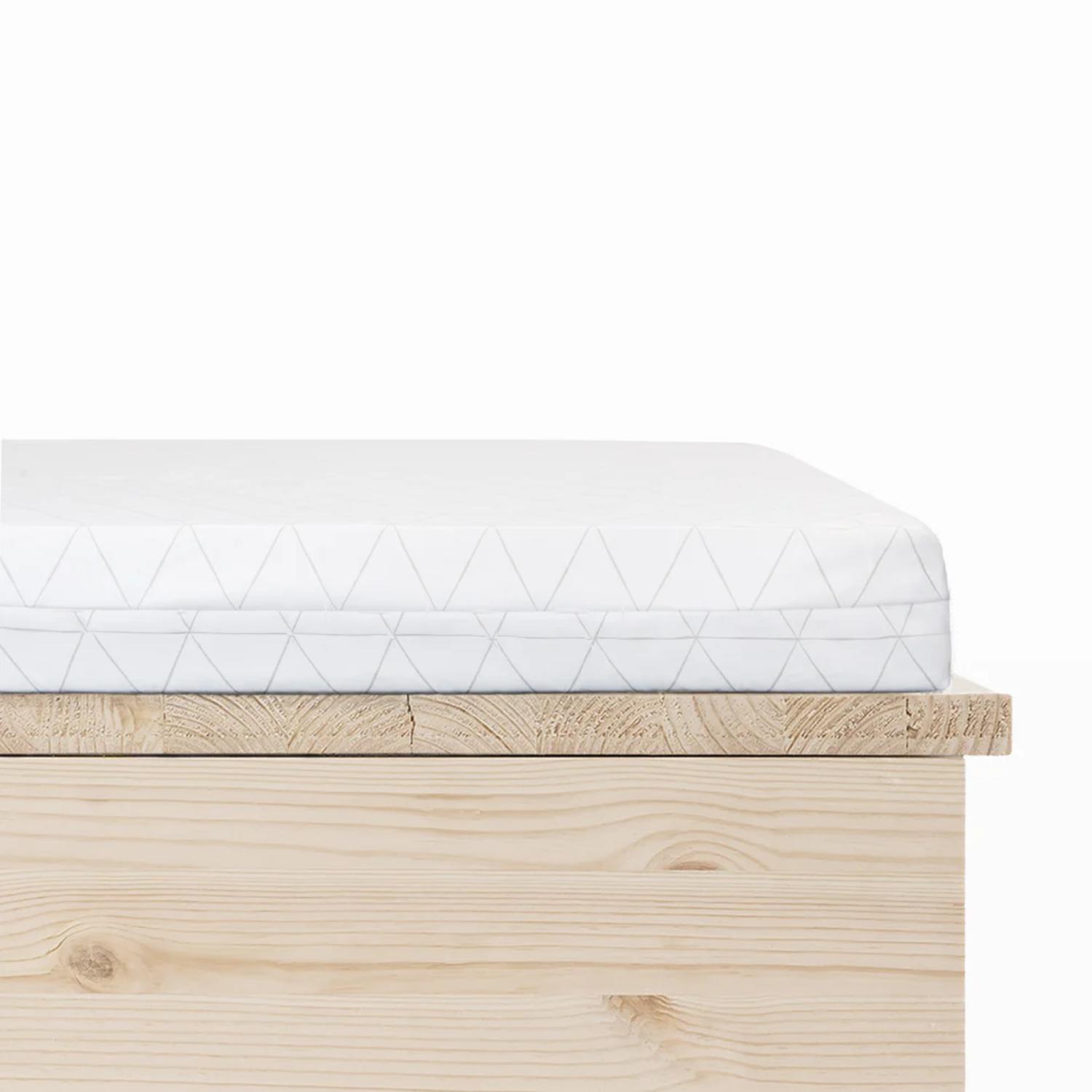 Lähikuva Vauvanpatja 60x120x5 original uusi kuosi -patjasta, jossa on valkoinen päällinen, jossa on hienovarainen kolmiokuvio, ja joka on asetettu vaaleanpuiseen puiseen sängynrunkoon valkoista taustaa vasten.