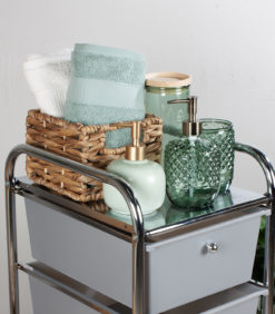 Korivaunun tummanharmaassa on kudottu kori, jossa on taiteltuja pyyhkeitä, kaksi lasista saippua-annostelijaa ja vihreä purkki, jossa on puukansi, kaikki siististi järjestetty valkoista seinää vasten.