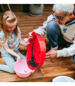 Iäkäs nainen kaataa marjoja punaisesta kulhosta valkoiseen kulhoon, jota nuori tyttö pitää kädessään, heidän istuessaan yhdessä ulkona puisilla portailla.