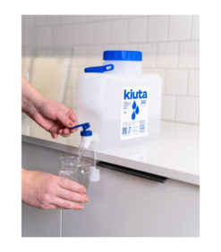 Henkilö annostelee vettä suuresta, suorakaiteen muotoisesta, valkoisesta muoviastiasta, jossa on merkintä kiuta, lasiin keittiön tason päällä, jossa on valkoinen laatoitettu välitila.