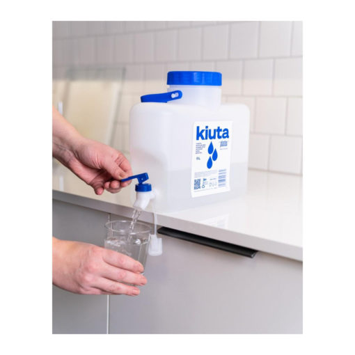 Henkilö annostelee vettä suuresta, suorakaiteen muotoisesta, valkoisesta muoviastiasta, jossa on merkintä kiuta, lasiin keittiön tason päällä, jossa on valkoinen laatoitettu välitila.