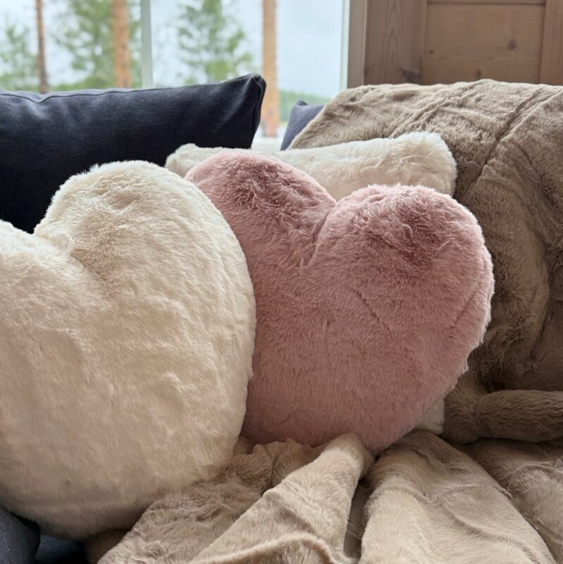 Sydämenmuotoiset valkoiset ja vaaleanpunaiset tyynyt lepäävät beigeä, pörröistä peittoa vasten, ja taustalla näkyy ikkuna, josta avautuu näkymä puille ja pilviselle taivaalle. Kodikas, lämmin ja kutsuva sisätilakuva.